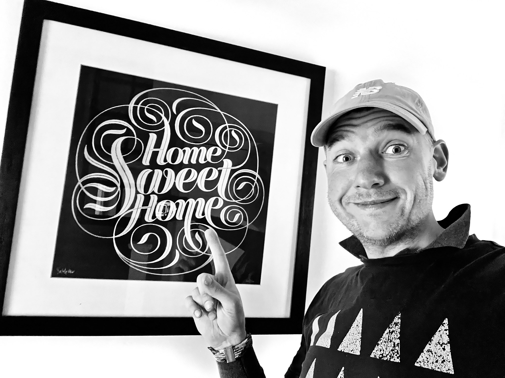 A photo of me smiling pointing to a framed hand written calligraphic print saying 'Home sweet home'